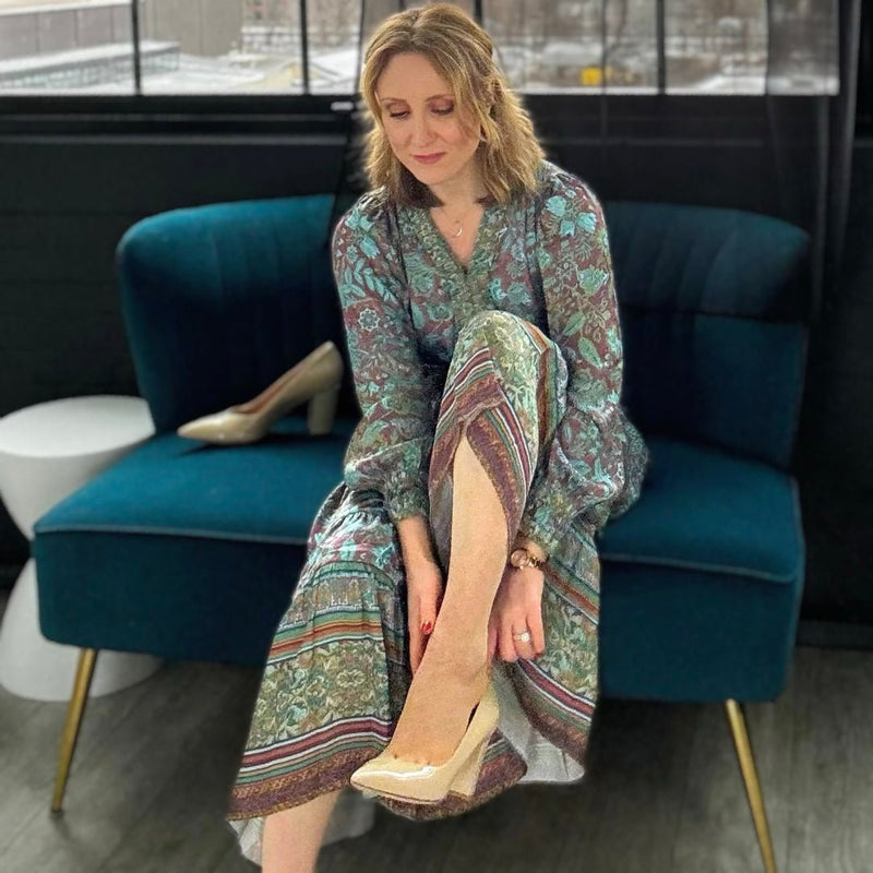 Woman in a patterned dress sitting on a blue couch in a room with a window, wearing Luna Selene nude pumps.
