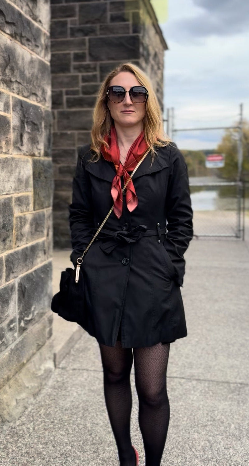 Woman wearing sunglasses, a black trench coat, and red Luna Selene silk scarf tied around her neck. 